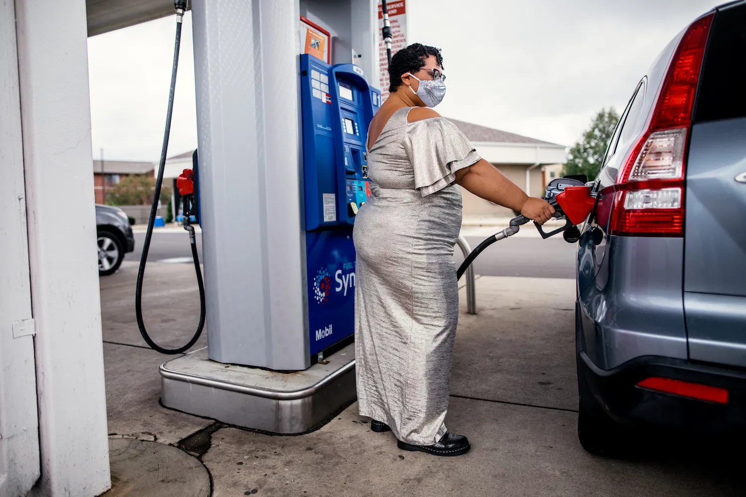 Professor Sami Schalk wearing a floor length silver dress, a silver sequin face mask, filling up a vehicle’s gas tank at a gas station.