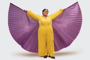 Sami Schalk wears a yellow jumpsuit and a shiny purple cape. Her arms are open to show the fullness of the cape. 
