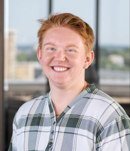 Portrait photo of Dean Dvorak standing in front of a window wearing a green and white plaid shirt.