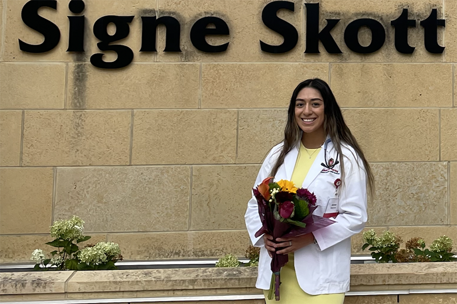 Aimee Melendez stands in front of Signe Skott Cooper Hall wearing a BSN white coat, a yellow dress, and holding a bouquet of flowers.