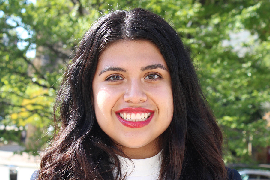 Alyssa Rodriguez smiles with green trees in the background. Alyssa has dark, flowing brown hair and wears red lipstick.