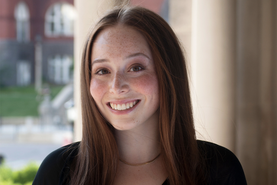 Ami Lieberman smiles at a window with Science Hall in the background. Ami has long brown hair and wears a black shirt.