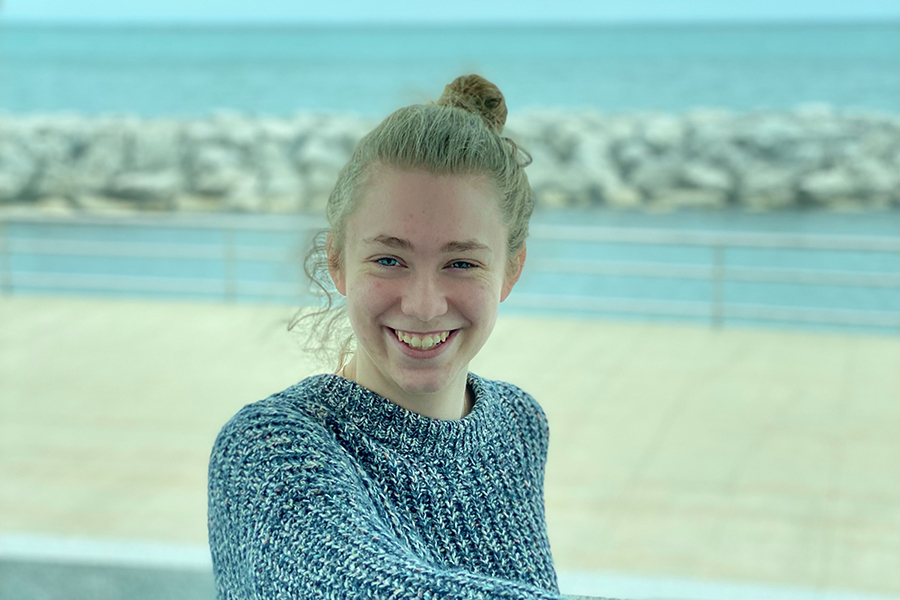 image of Annika Zuverink sitting outside in front of sand and water with blonde/brown hair pulled back into a bun smiling at the camera wearing a blue/grey sweater.
