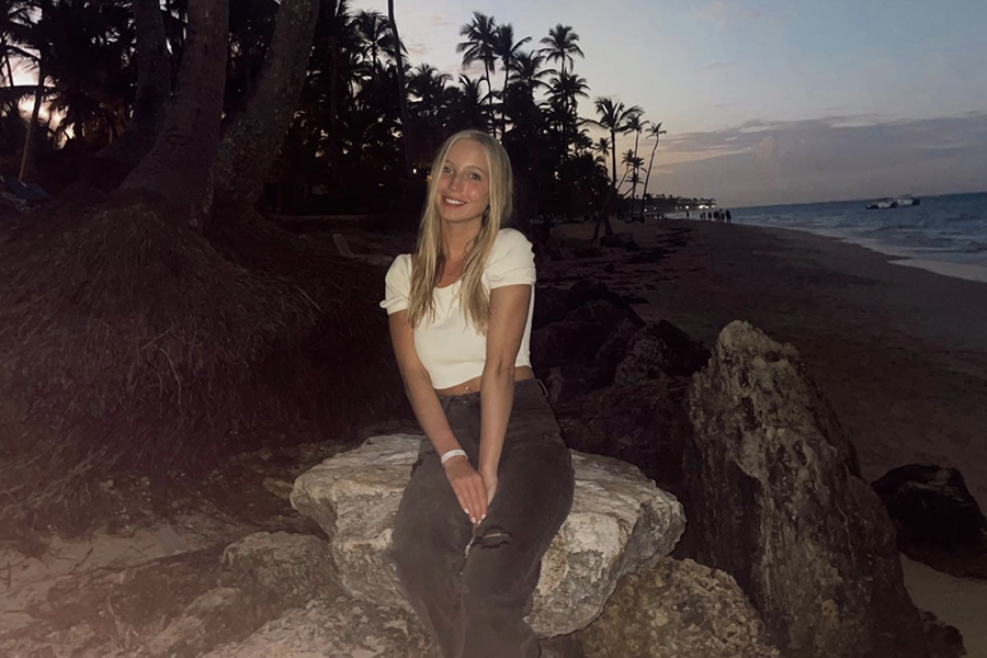 Ashlyn Friedrichs poses in front of rocks, palm trees, and the beach. Ashlyn is wearing a light, short sleeve short and dark pants.