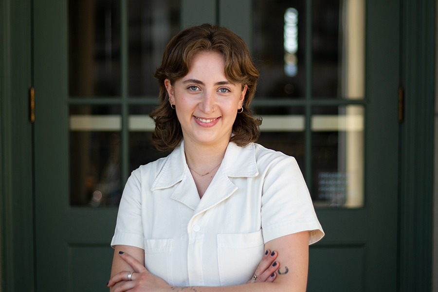 Caitlyn Halfon stands with arms crossed and wearing an ivory short-sleeved shirt in front a glass-paned dark green door. Carolyn has shoulder-length brown hair.