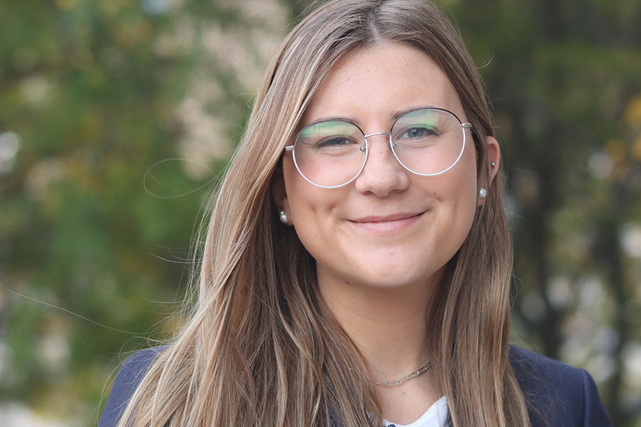 Erin Kennon smiles in front of a leafy background. Erin has long blond hair, wears glasses, and wears a white shirt and a blue jacket.