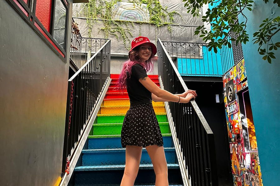image of Erin Wruk standing in front of rainbow-colored steps turning towards the camera and smiling wearing a dress and smiling