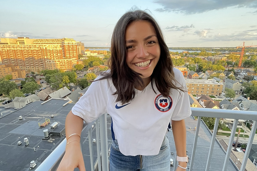 Francisca Ibanez stands on a balcony with a view of an urban landscape behind her. Francisca wears a white shirt and jeans and has long brown hair.
