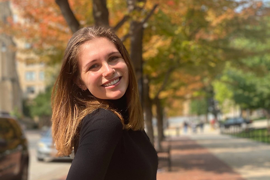Justice smiles with Library Mall in the background and trees with orange-green leaves. Justice has long brown hair and wears a black shirt.