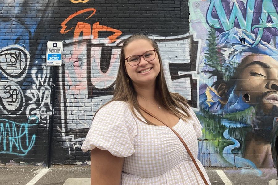 Karsyn Garber stands in front of a mural. Karsyn has long brown hair, is wearing glasses, and a white short.