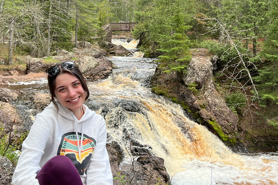 image of Olivia Van Hammond sitting outside near a waterfall wearing a white sweatshirt smiling at the camera with brown hair pulled back and sunglasses on top of head.