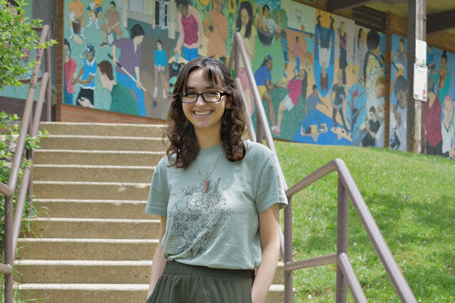 Rachel Litchman stands in front of concrete steps with a colorful mural in the background. Rachel is smiling and is wearing a pale green t-shirt and darker green shorts.