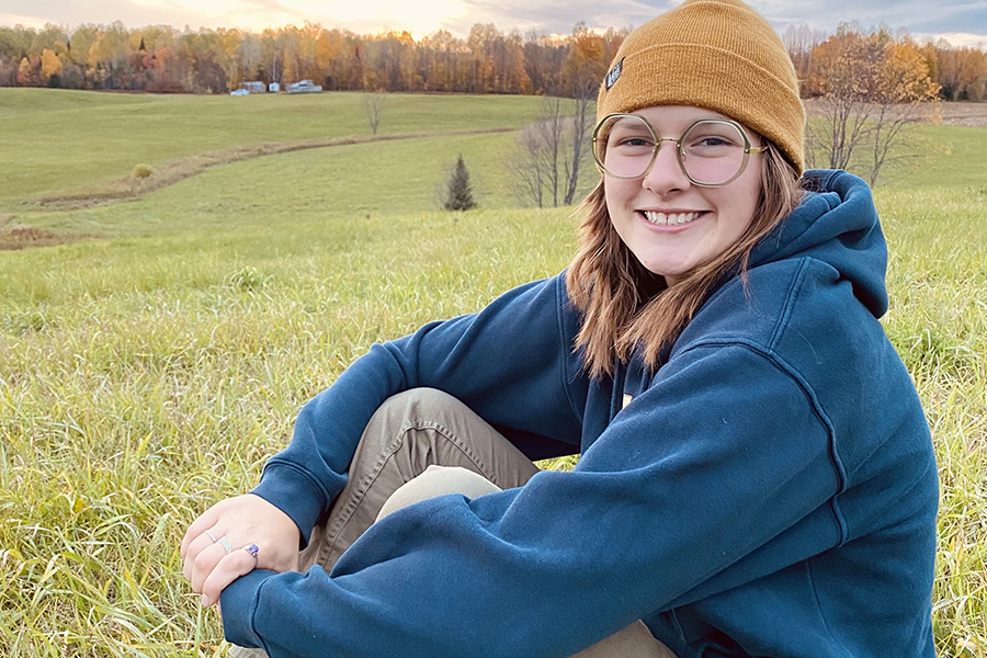 Reilly Coon is seated in a grassy field with fall foliage in the distance. Reilly is smiling, wearing a yellow hat and a blue hoodie.