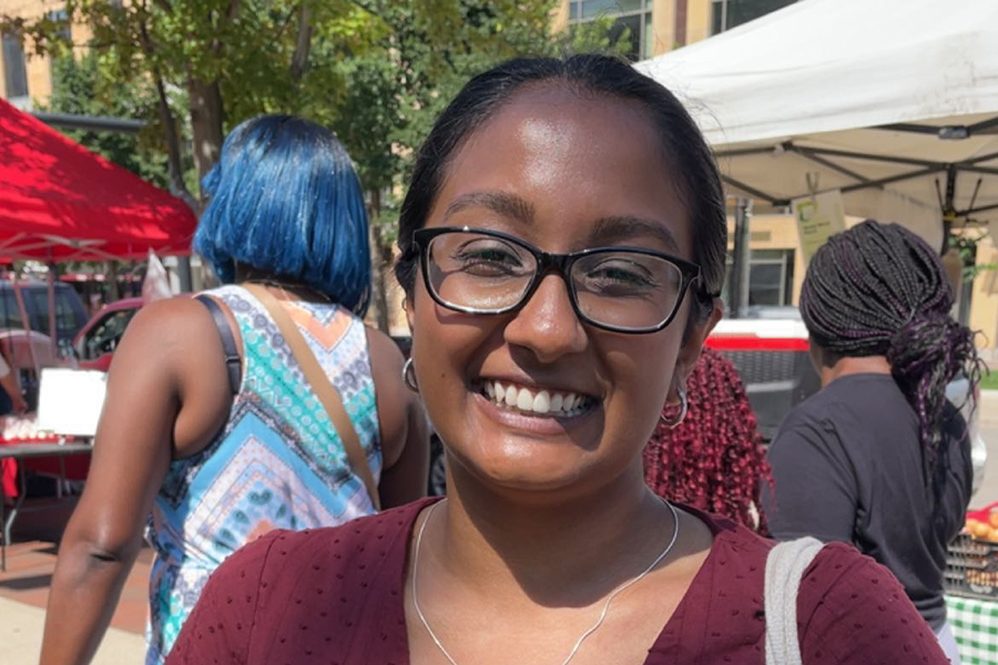Roshnie Rupnarain smiles wearing a burgundy shirt and her hair pulled back with market stalls in the background.