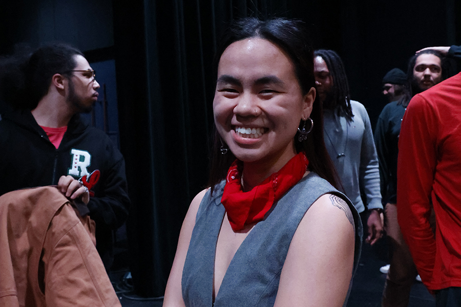 image of Samantha Starks standing and smiling at the camera. Samantha has long black hair pulled back, is wearing a red handkerchief around their neck and a grey/blue sleeveless top. They are smiling at the camera.