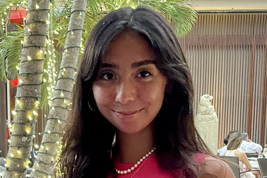 Sofia Irigoyen Barrientos smiles in front of trees wrapped with lights. Socia wears a bright pink dress and a pearl necklace.