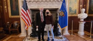 Alanna Goldstein with WI Governor Tony Evers standing in front of a decorative fireplace framed by the U.S. flag and the WI flag.