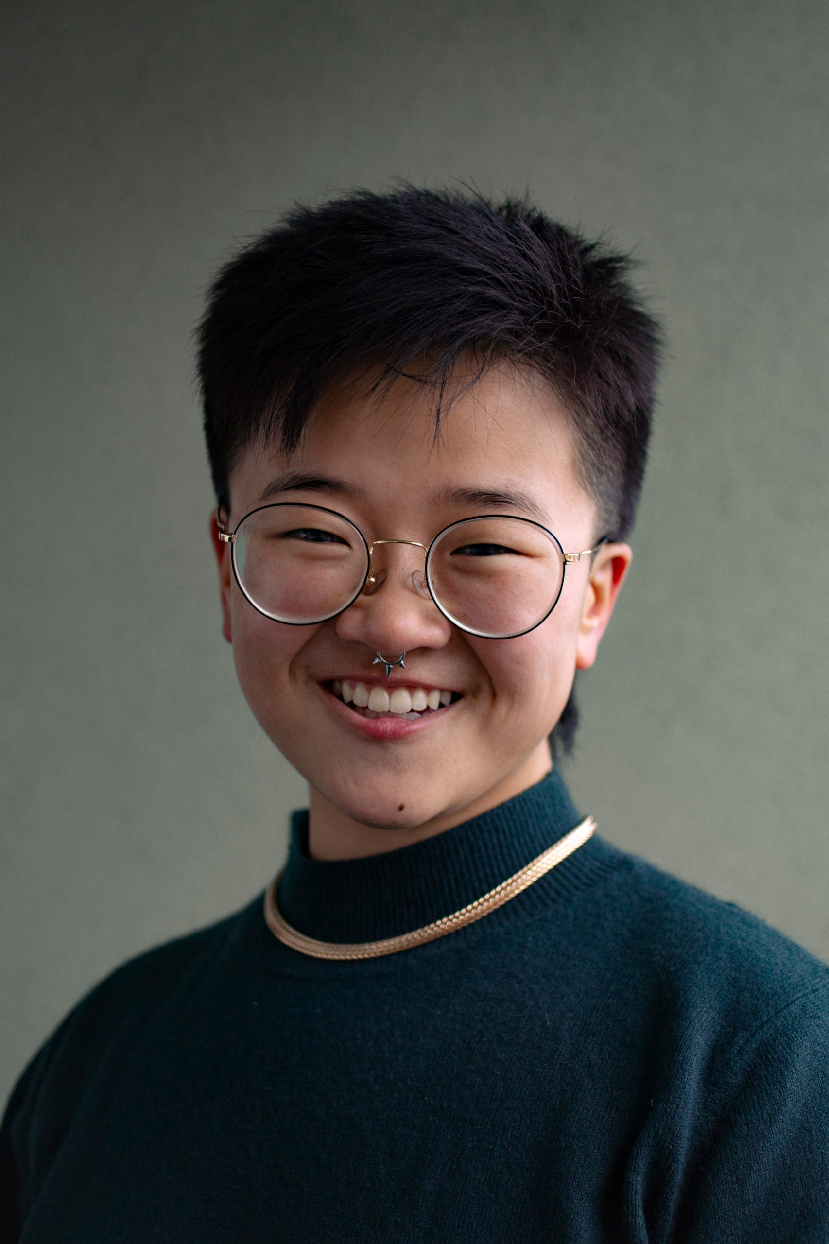 image of Koda Parker sitting and smiling at the camera with short nearly buzzed black hair and big circular glasses wearing a dark teal turtleneck and gold necklace.