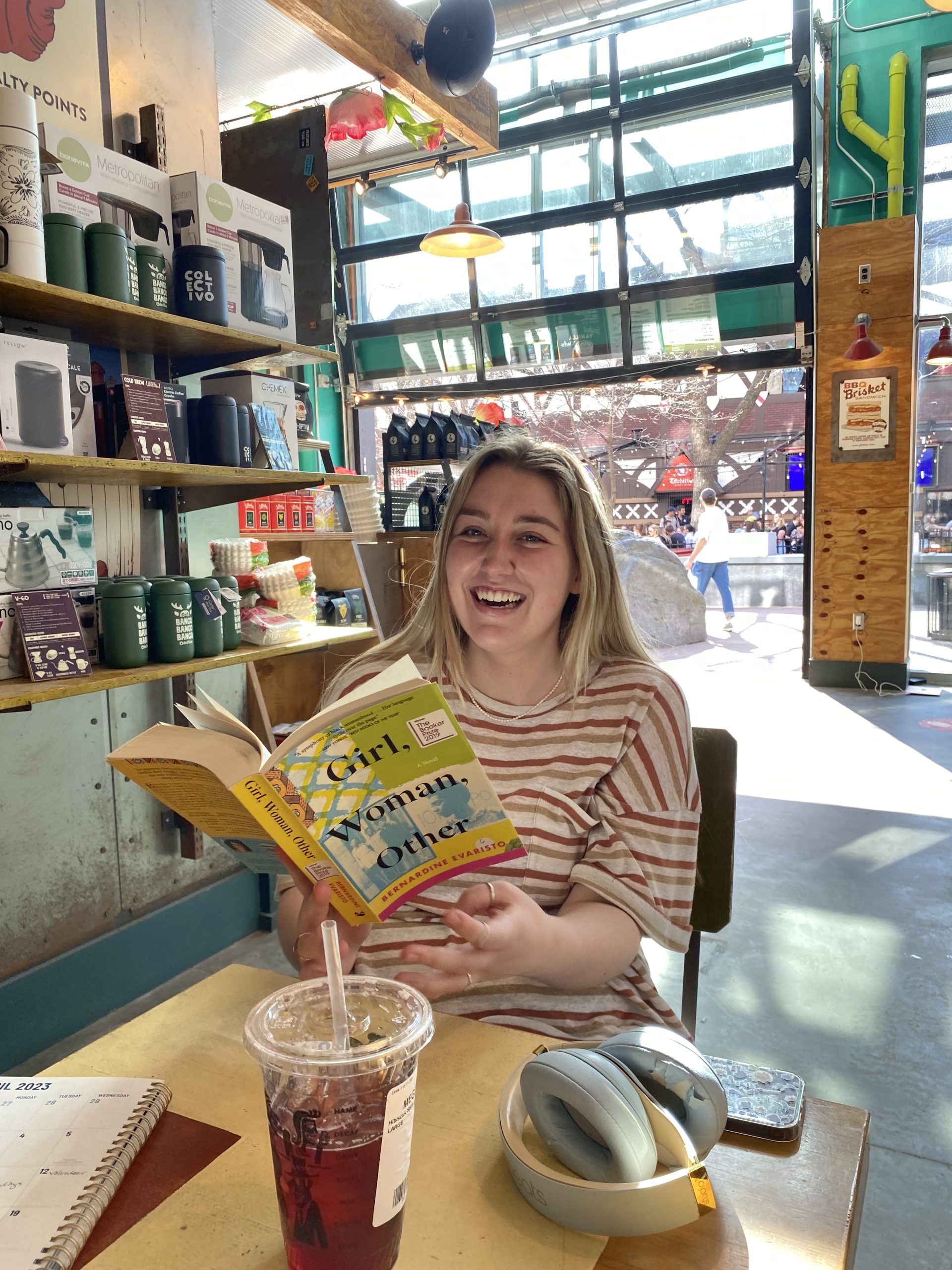 Megan Gahart is sitting in a cafe holding a book and smiling. Megan has long blond hair and is wearing a horizontal striped shirt. In the background, the doors of the café are open to the outdoors.