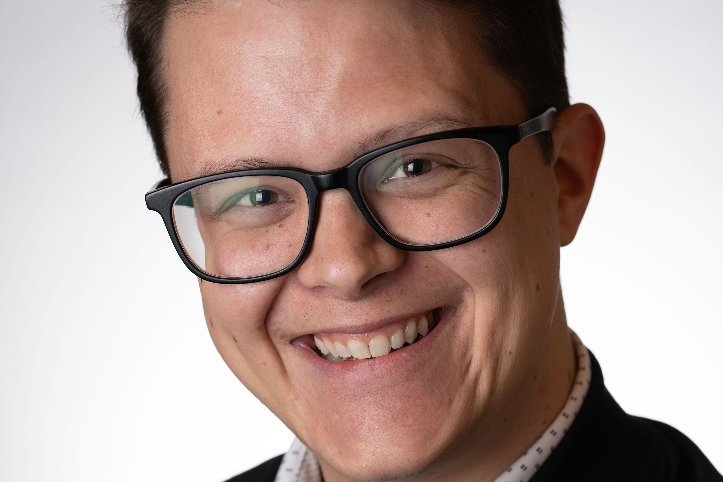Orion Risk smiles in this portrait photo wearing glasses, a collared shirt, and a dark jacket.