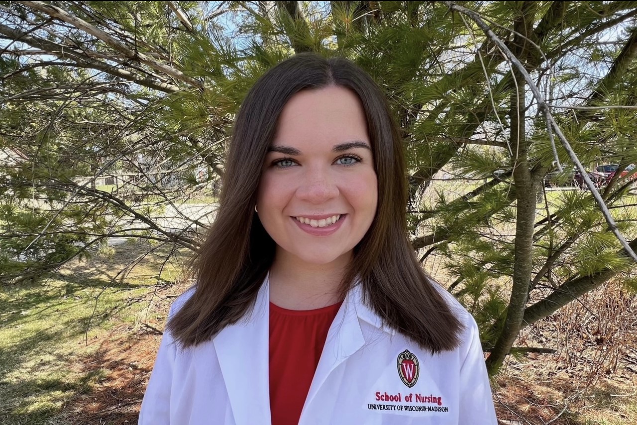 Paige Henry smiles in this portrait wearing a BSN white coat in front of a pine tree.