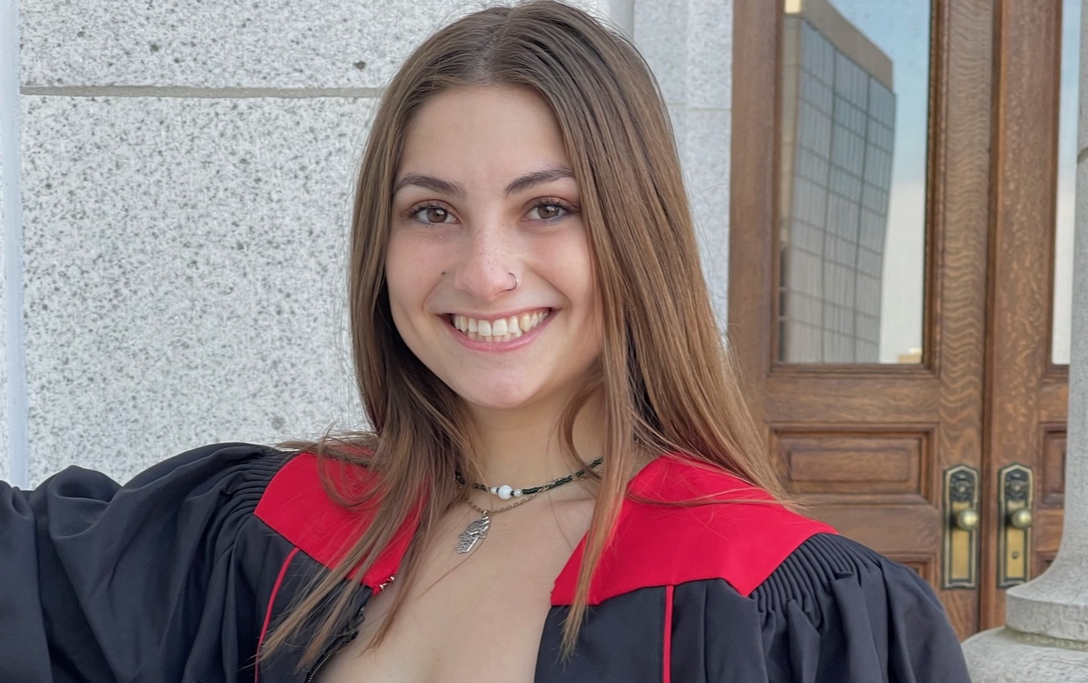 Rachel Mirviss smiles wearing a red and black graduation gown in front of a stone wall and glass-paned wooden doors with brass handles.