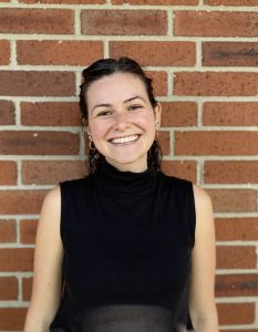 Image of Visiting Assistant Professor Molly M. Henderson. She stands against a warm brown brick wall. She has dark brown hair and wears a black sleeveless shirt.