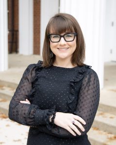 Photo of Hailee Yoshizaki-Gibbons wearing a black shirt, black framed glasses, a smile, and crossing her arms in front of steps.